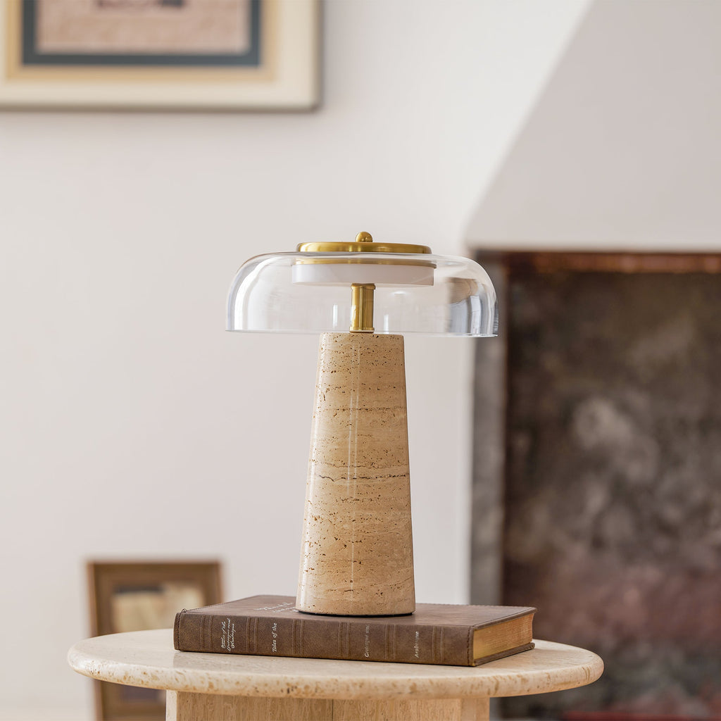 Clérachic table lamp with a clear glass dome, standing on a hardback book on a round travertine side table in a softly lit living room.