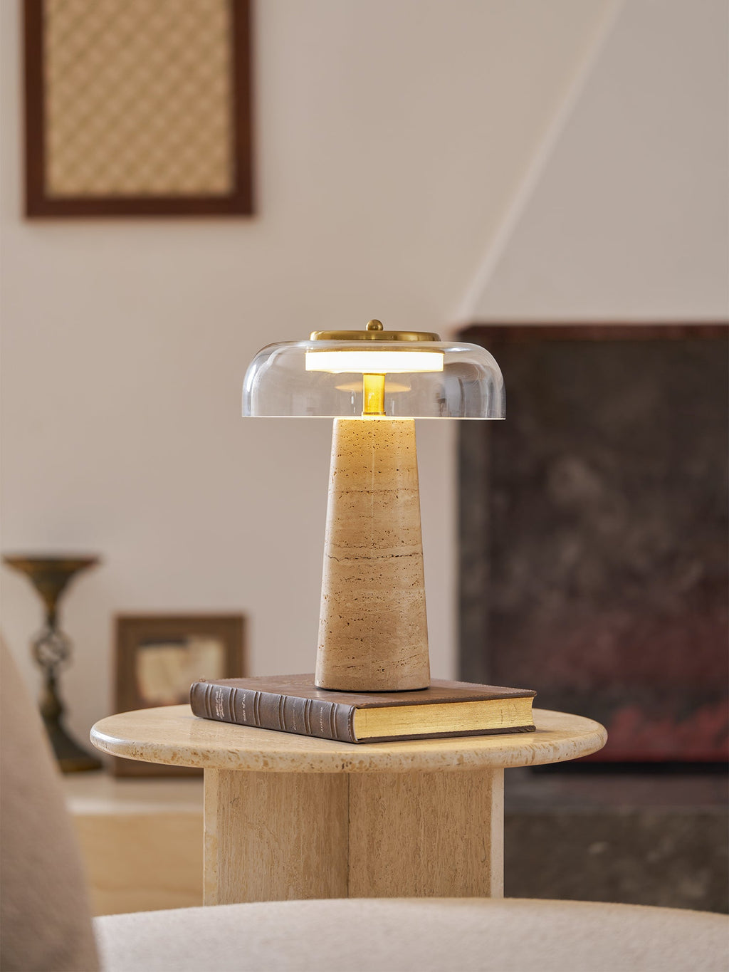 Clérachic table lamp lit on a book atop a round travertine table, with a framed picture and small vase behind it.