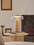 Clérachic table lamp lit on a book atop a round travertine table, with a framed picture and small vase behind it.