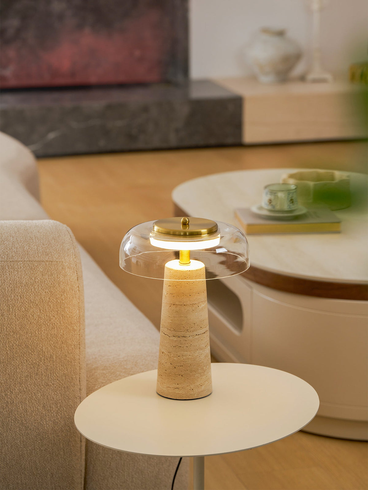 Lit Clérachic lamp on a round white table, the warm light glowing under the clear dome and up the travertine base.