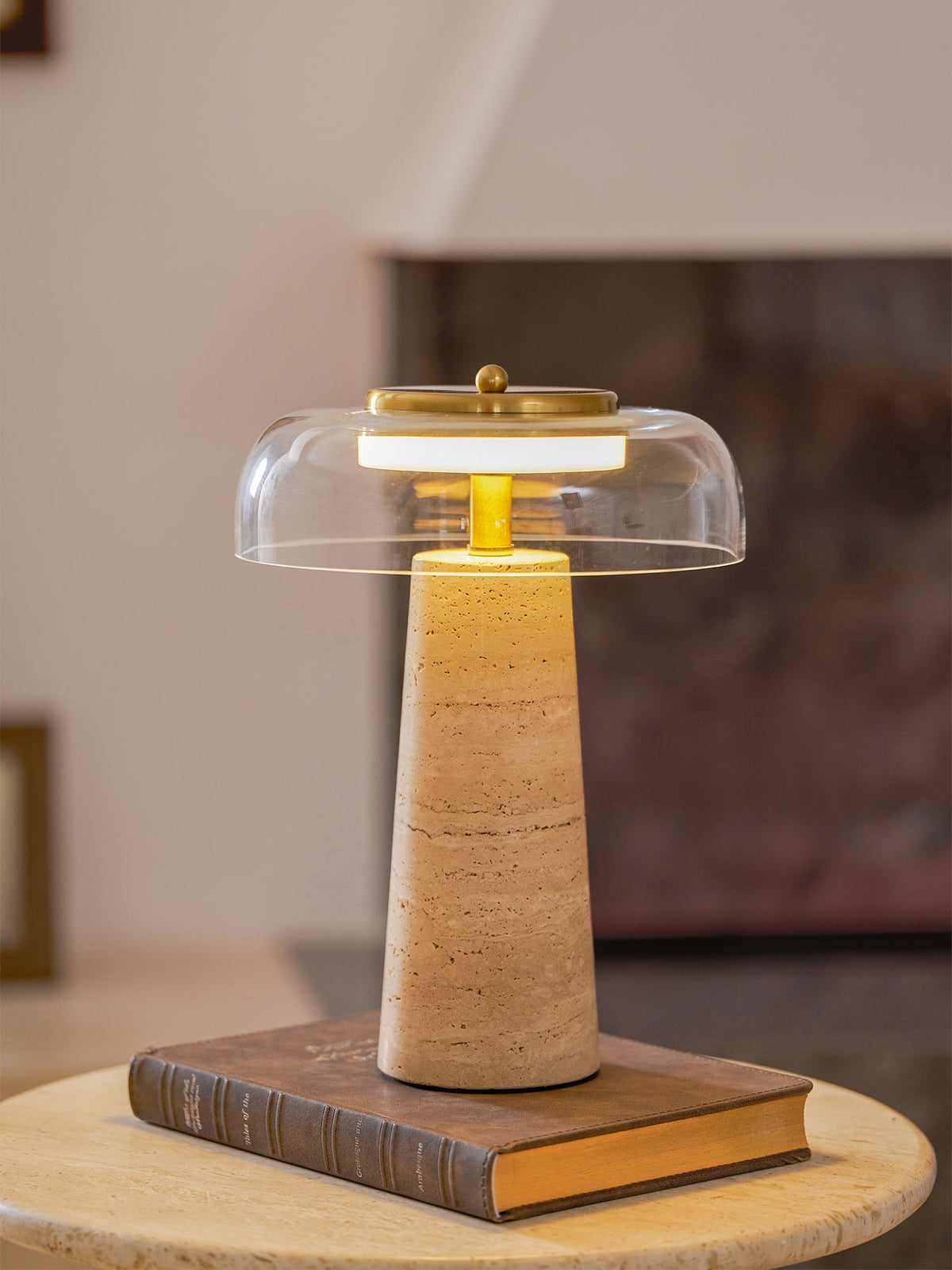 Low angle on this Clérachic table lamp lit on a leather-bound book, its tall travertine base filling the frame.