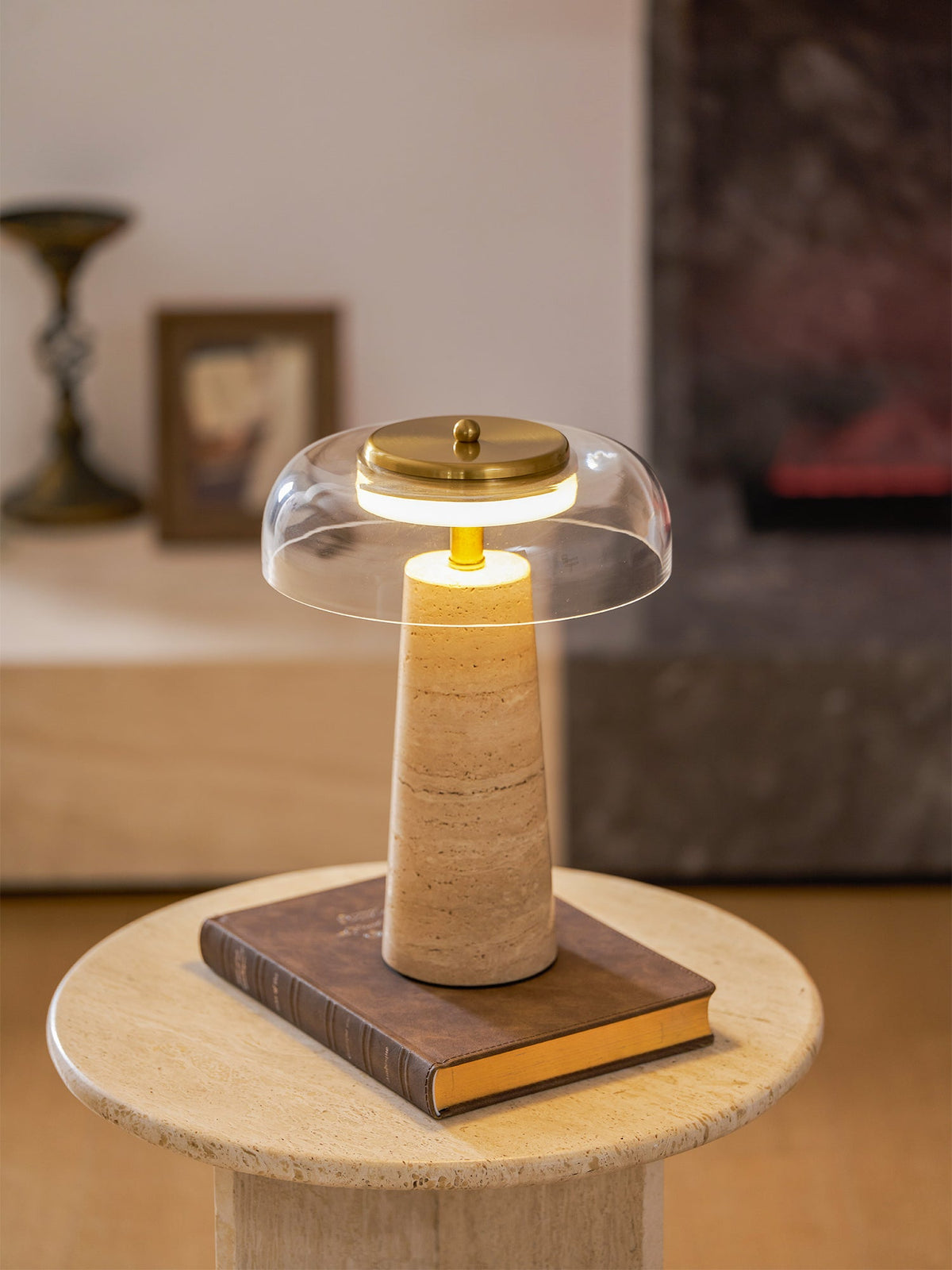 Clérachic table lamp glowing on a hardback book over a round travertine side table, with a framed picture beyond.