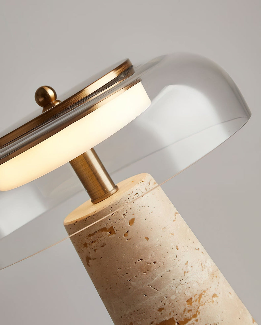 Angled close view of the brass stem and clear glass dome on this Clérachic table lamp, where the travertine base meets the stem.