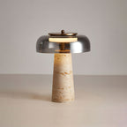 This Clérachic table lamp with a smoky grey dome, lit, its travertine base showing orange-brown veining against a pale grey wall.