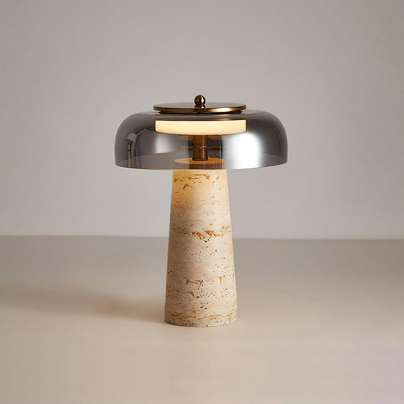 This Clérachic table lamp with a smoky grey dome, lit, its travertine base showing orange-brown veining against a pale grey wall.