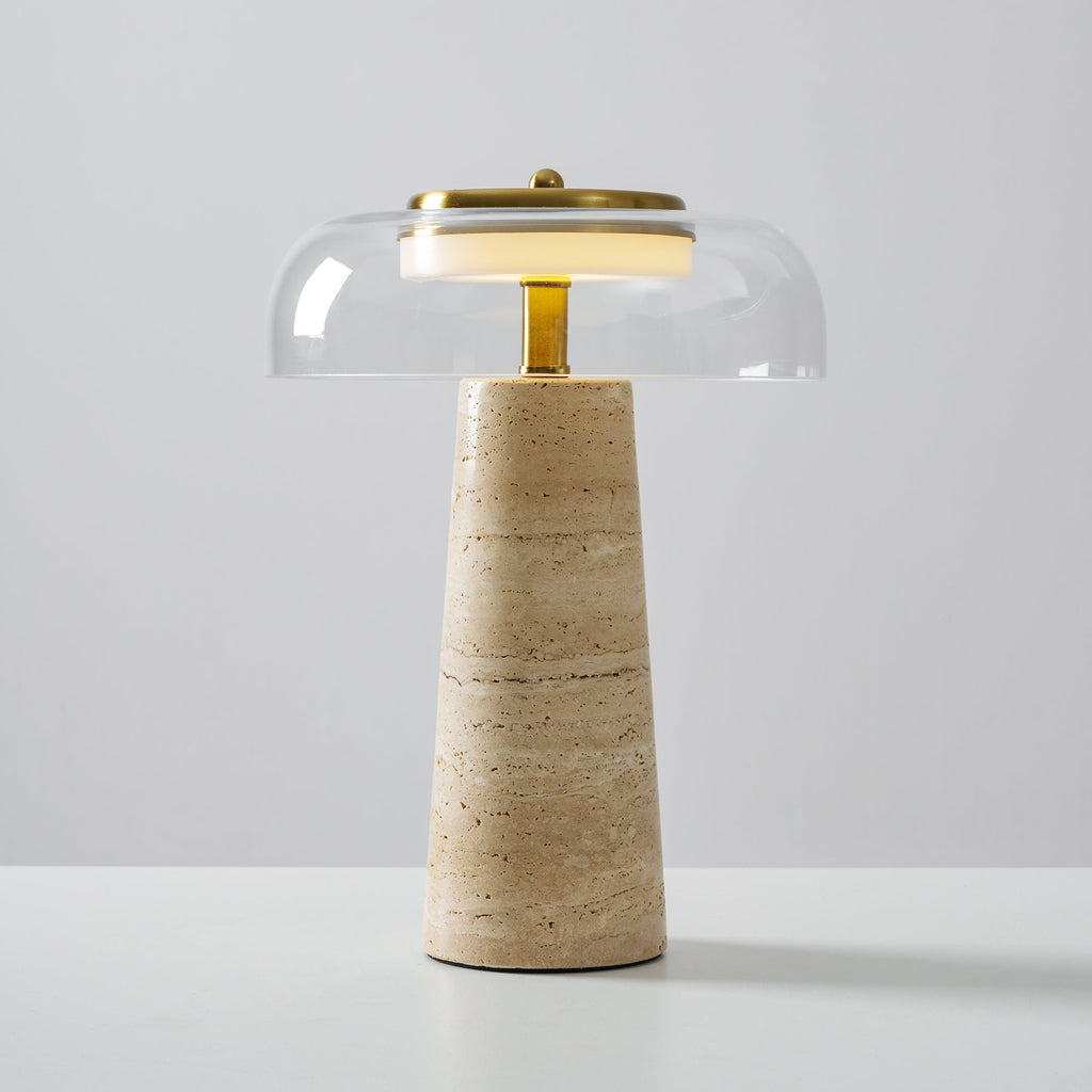 Clérachic table lamp with a clear glass dome and brass stem, its banded beige travertine base standing on a white surface.