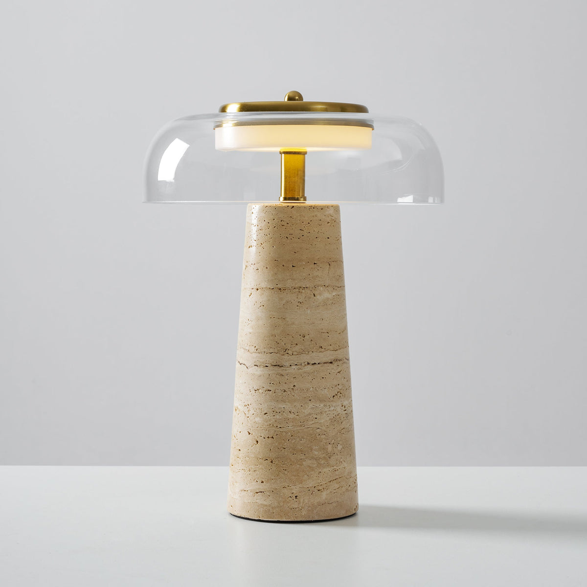 Clérachic table lamp with a clear glass dome and brass stem, its banded beige travertine base standing on a white surface.
