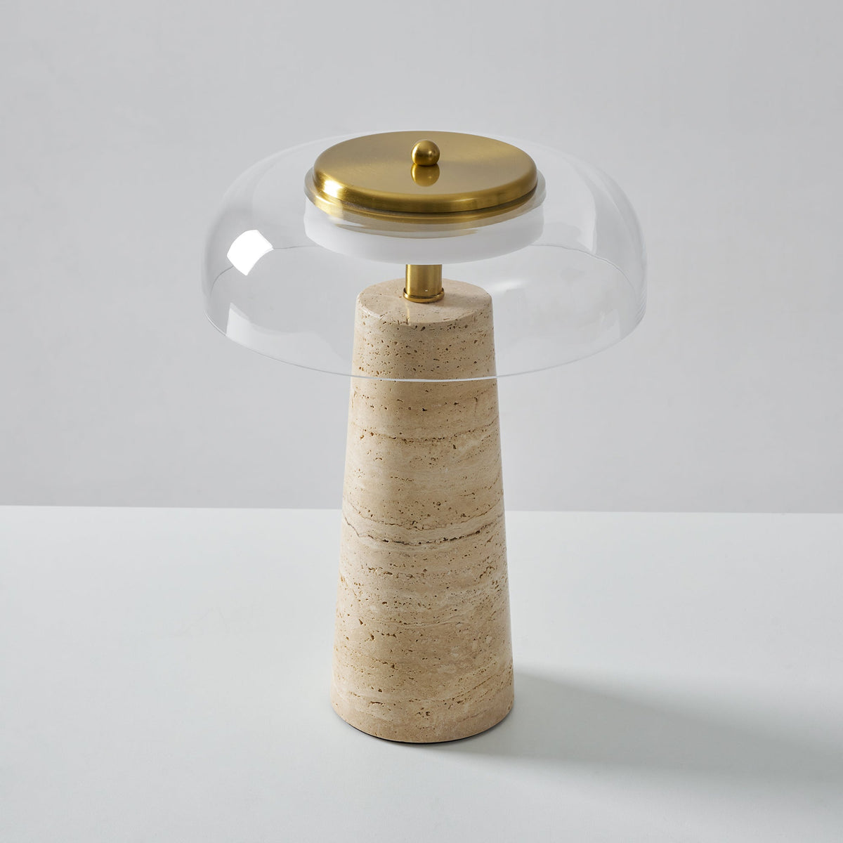 Slightly higher view of this Clérachic table lamp, a brass cap crowning a clear glass dome above the travertine base.