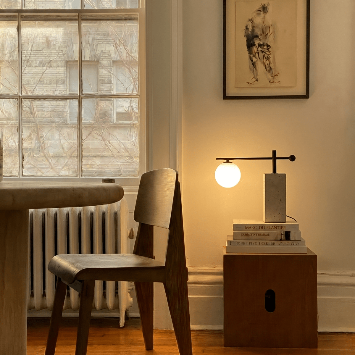 This Clérachic table lamp glows on a wooden side table stacked with books beside a woven chair.