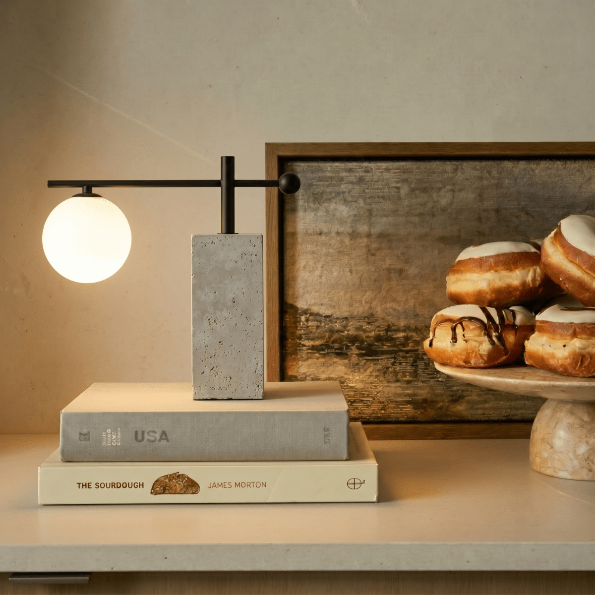 This Clérachic table lamp glows atop a stack of books on a console set with pastries nearby.