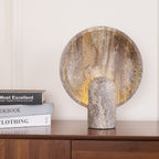 A Clérachic travertine lamp glowing on a walnut sideboard, beside a short stack of books against a cream wall.