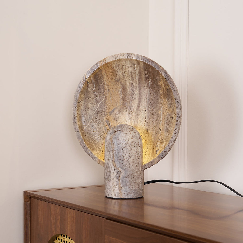 Round stone disc lamp from Clérachic standing at the corner of a walnut cabinet, its black cord trailing off to the right.