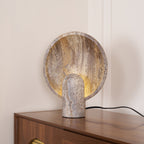 Round stone disc lamp from Clérachic standing at the corner of a walnut cabinet, its black cord trailing off to the right.