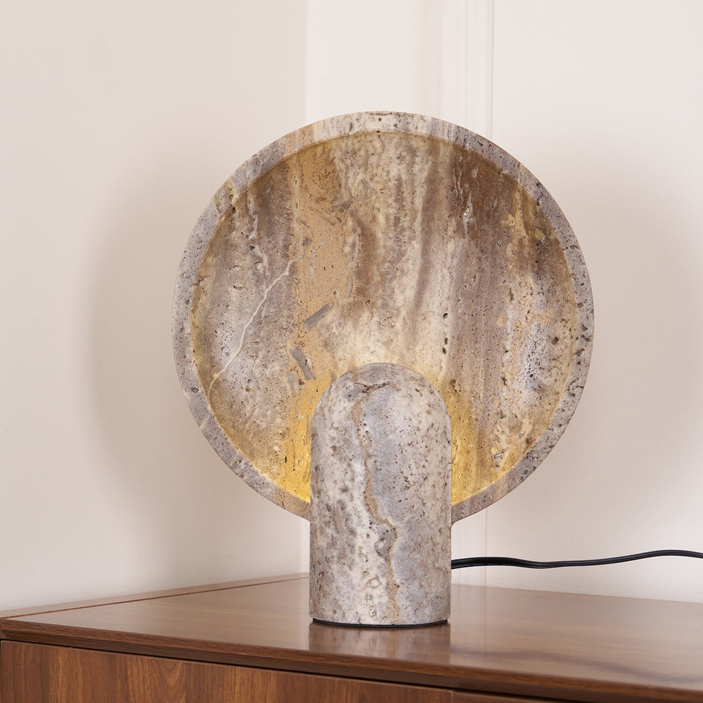 Close, slightly raised view of the Clérachic travertine lamp, its banded brown and cream disc lit warmly from behind the stone stem.