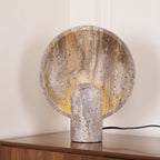 Close, slightly raised view of the Clérachic travertine lamp, its banded brown and cream disc lit warmly from behind the stone stem.