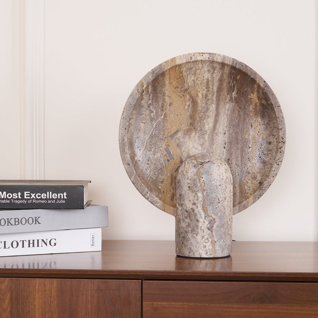 The brown travertine lamp from Clérachic on a wooden sideboard next to stacked books, the disc catching a soft amber glow.