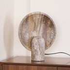 Three-quarter view of a Clérachic stone lamp on a walnut cabinet, showing the veined, pitted face of the rounded disc.