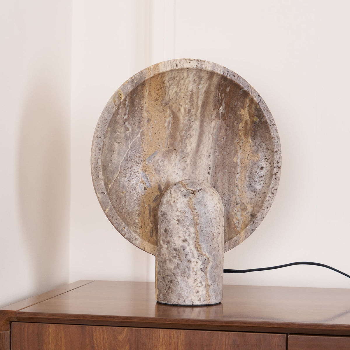 Three-quarter view of a Clérachic stone lamp on a walnut cabinet, showing the veined, pitted face of the rounded disc.