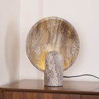 Stone table lamp from Clérachic on a dark wood chest, the lit disc standing behind a short, rounded travertine post.