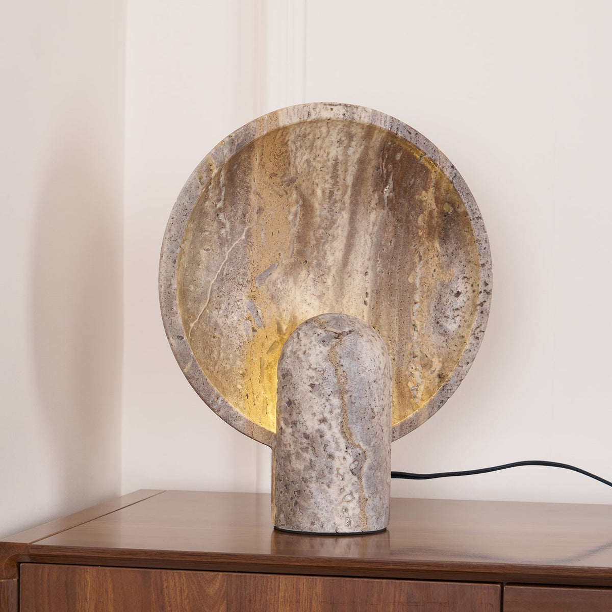 Stone table lamp from Clérachic on a dark wood chest, the lit disc standing behind a short, rounded travertine post.