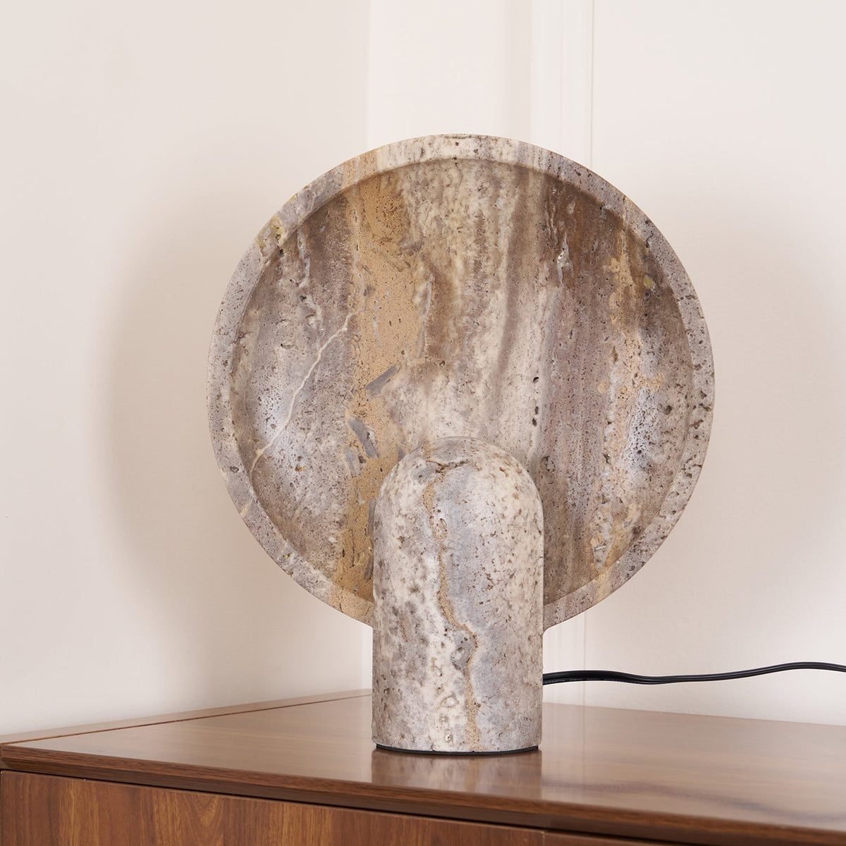 A Clérachic travertine lamp in a corner of a walnut sideboard, the disc glowing amber with the black cord looping behind.