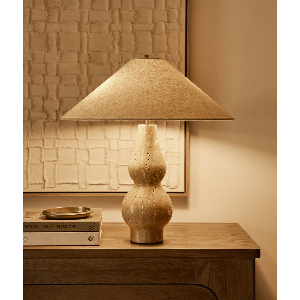 On a wooden chest beside a textured cream wall panel, this lit Clérachic table lamp shows a warm linen shade over a pitted travertine base.