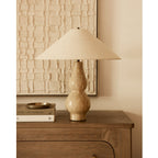 With its shade softly lit, this Clérachic table lamp stands on a wooden chest in front of a textured cream artwork and a stack of books.
