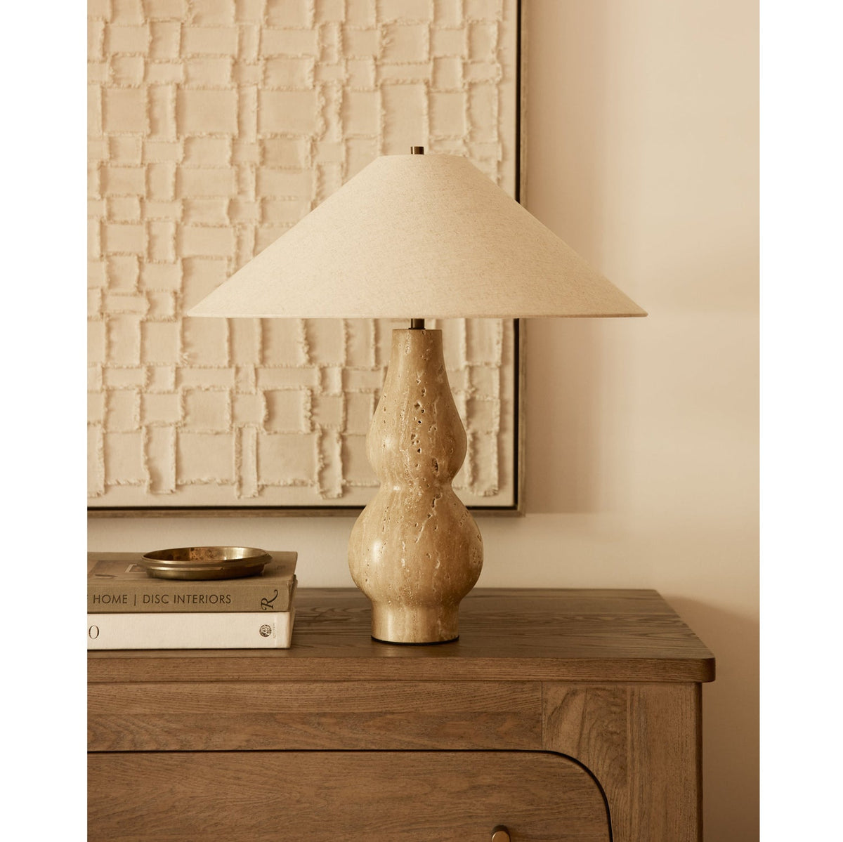 With its shade softly lit, this Clérachic table lamp stands on a wooden chest in front of a textured cream artwork and a stack of books.