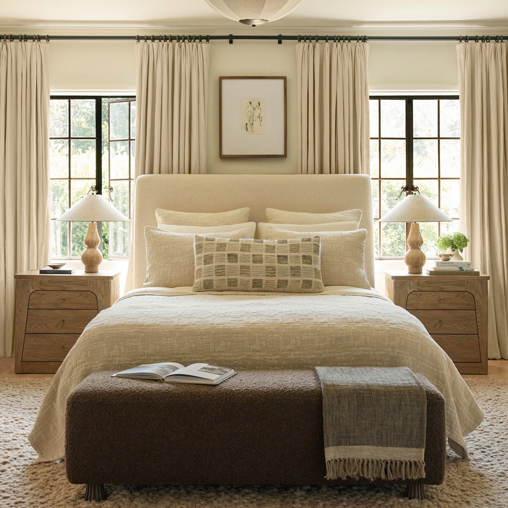 Two Clérachic table lamps with white linen shades sit on either bedside chest, on both sides of a cream bed in a bright, curtained room.