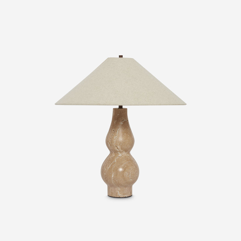 Clérachic table lamp on a light grey backdrop, a beige linen shade above a sand-coloured travertine base with dark metal detail at the foot.