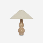 Clérachic table lamp on a light grey backdrop, a beige linen shade above a sand-coloured travertine base with dark metal detail at the foot.