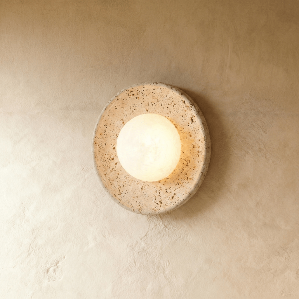 Clérachic round travertine wall light with a lit frosted white globe at its centre, set against a soft limewashed wall.