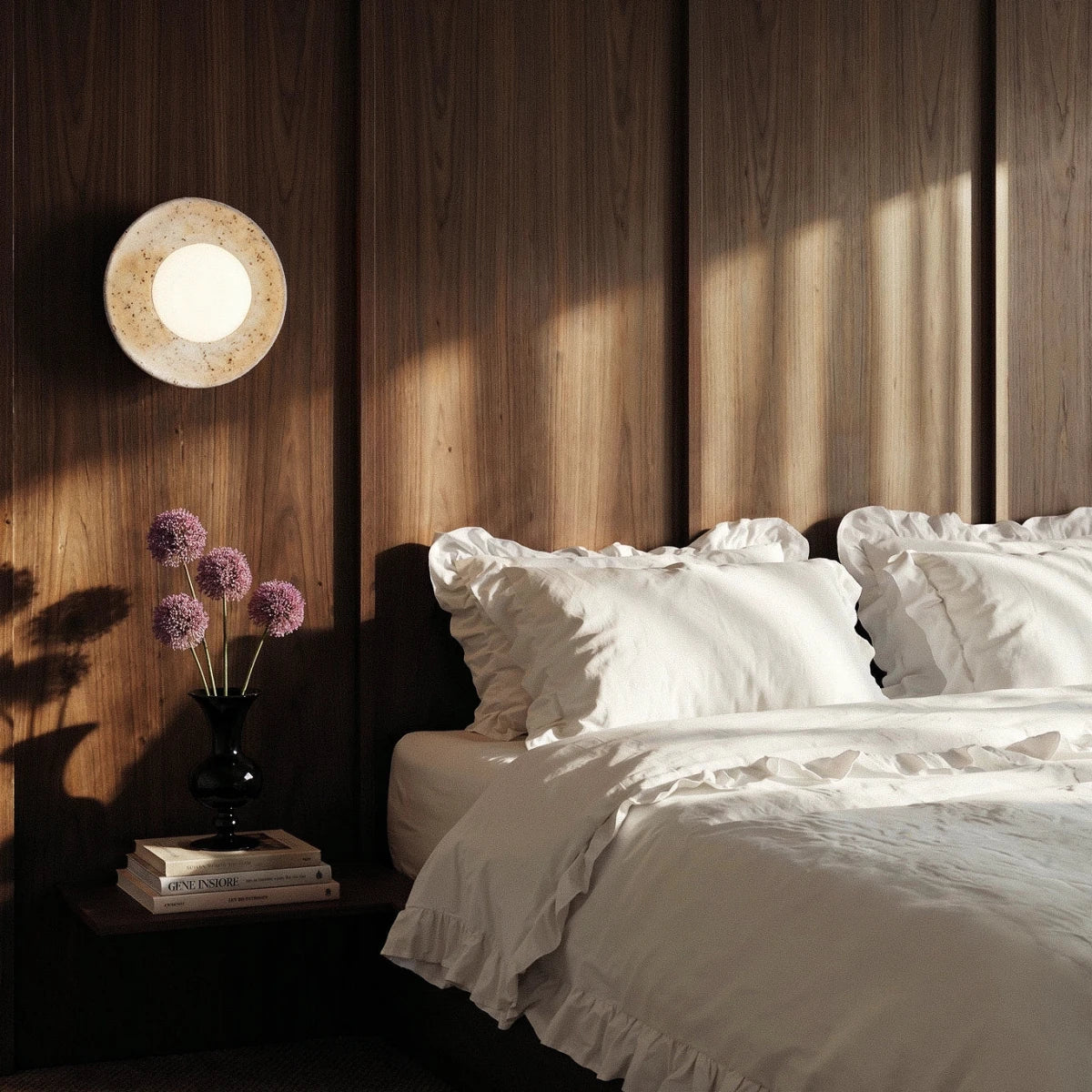 Clérachic round travertine wall light glowing above a bedside table with books and alliums, against walnut panelling.