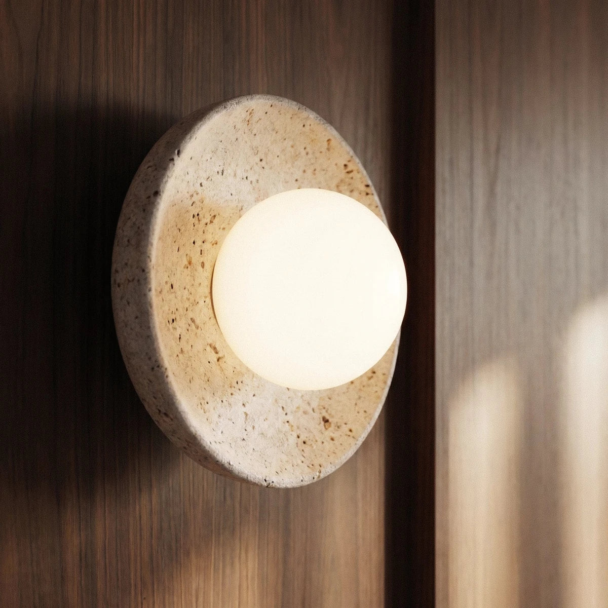Clérachic round travertine wall light with a glowing opal globe, seen at an angle against warm walnut panelling.