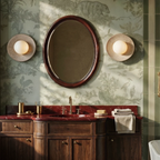 Clérachic pair of round travertine wall lights flanking an oval mirror above a bathroom vanity, on sage toile wallpaper.