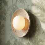 Clérachic travertine wall light with a glowing opal globe, seen at an angle against sage green botanical wallpaper.