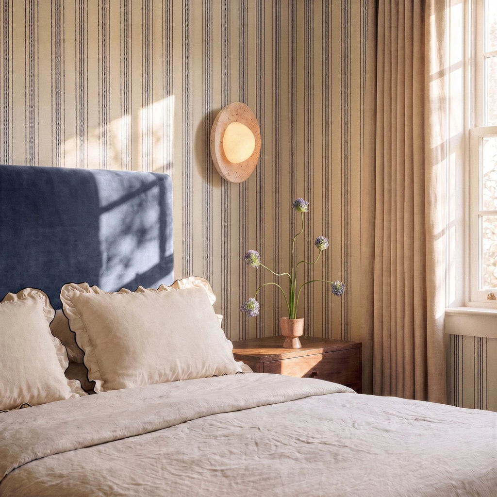 Clérachic round travertine wall light glowing above a bedside table with flowers, on striped wallpaper beside a blue headboard.