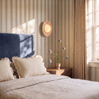 Clérachic round travertine wall light glowing above a bedside table with flowers, on striped wallpaper beside a blue headboard.