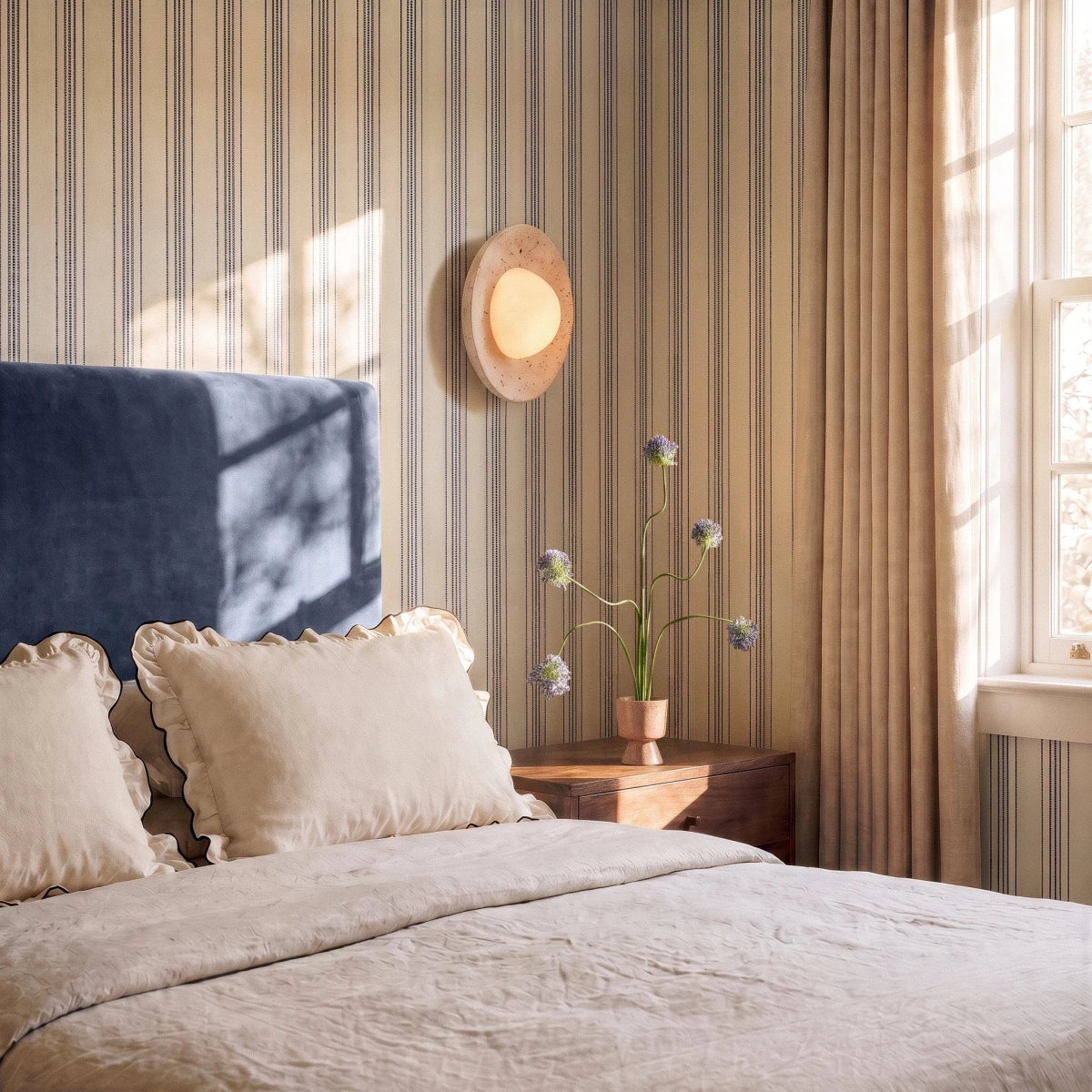 Clérachic round travertine wall light glowing above a bedside table with flowers, on striped wallpaper beside a blue headboard.