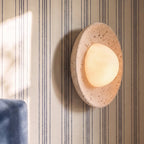 Clérachic travertine wall light with a softly lit, pebble-shaped opal globe, seen close up on striped cream wallpaper.