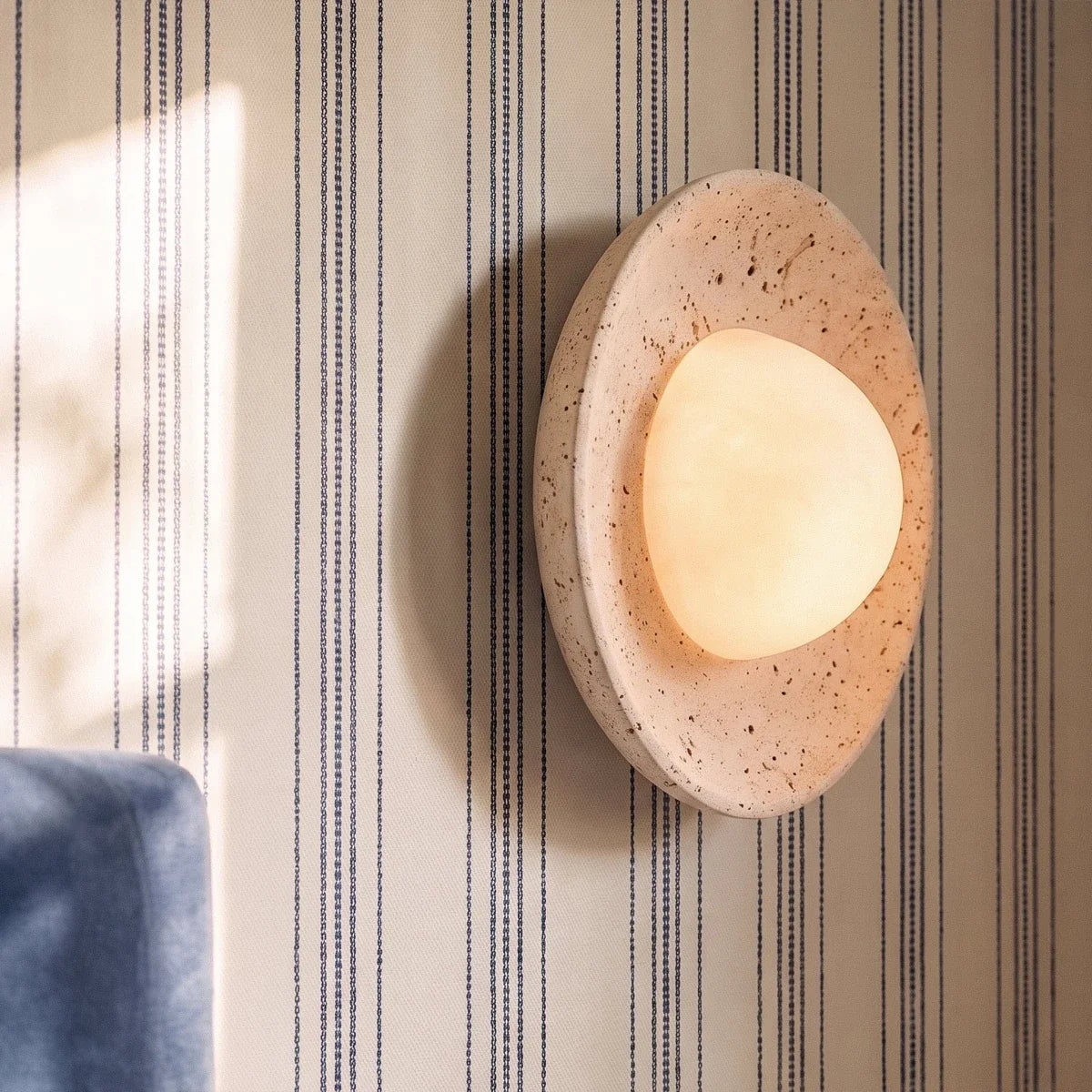 Clérachic travertine wall light with a softly lit, pebble-shaped opal globe, seen close up on striped cream wallpaper.