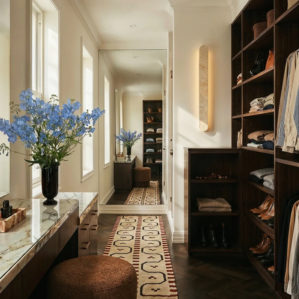 Clérachic cream travertine wall light glowing warmly in a dressing room with dark wood shelving, a marble vanity and a runner.