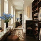 Clérachic cream travertine wall light glowing warmly in a dressing room with dark wood shelving, a marble vanity and a runner.