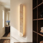 Clérachic travertine wall light close up, showing the pitted cream stone and a soft glow behind it beside dark wood shelves.