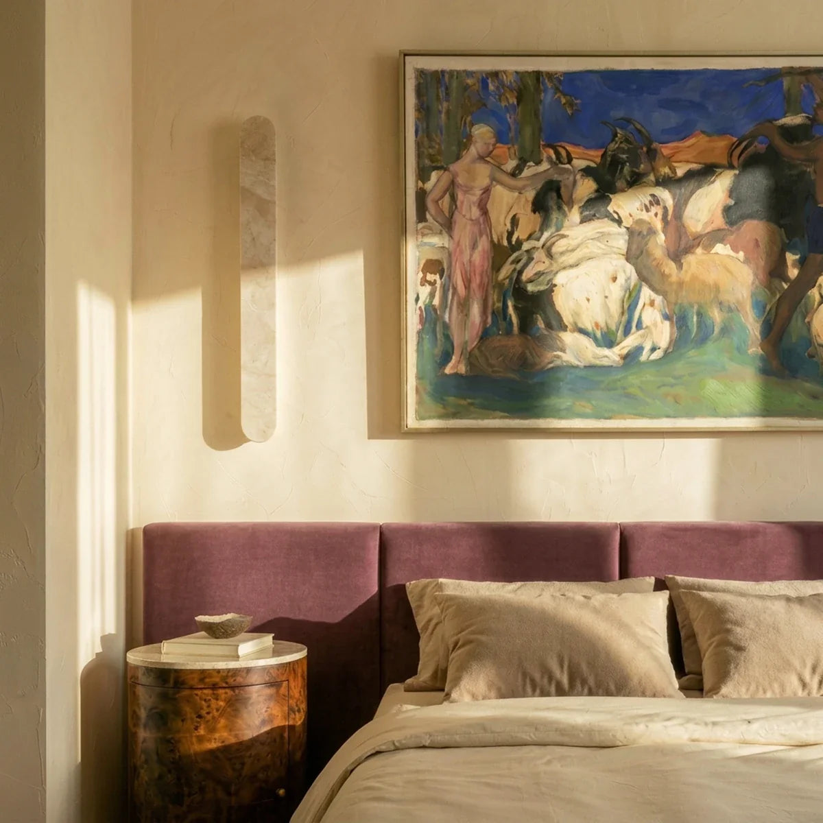 Clérachic cream travertine wall light mounted beside a framed painting above a plum headboard, with sunlight across the wall.