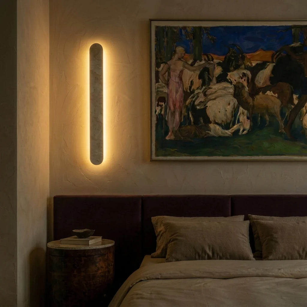 Clérachic travertine wall light lit at dusk beside a framed painting, giving the bedroom wall a warm halo behind the stone.