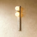Clérachic vertical travertine wall lamp with two glowing frosted glass globes, mounted on a warm limewashed plaster wall.