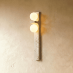 Clérachic vertical travertine wall lamp with two glowing frosted glass globes, mounted on a warm limewashed plaster wall.
