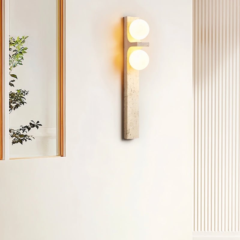 Clérachic travertine wall lamp with two glowing globes beside a tall mirror and a fluted panel in a bright, calm room.