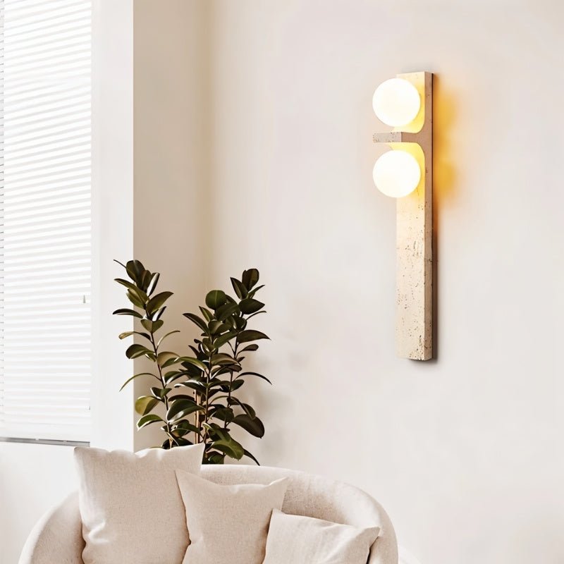 Clérachic travertine wall lamp lit beside a sofa and a leafy plant, giving a soft amber glow on a pale wall.